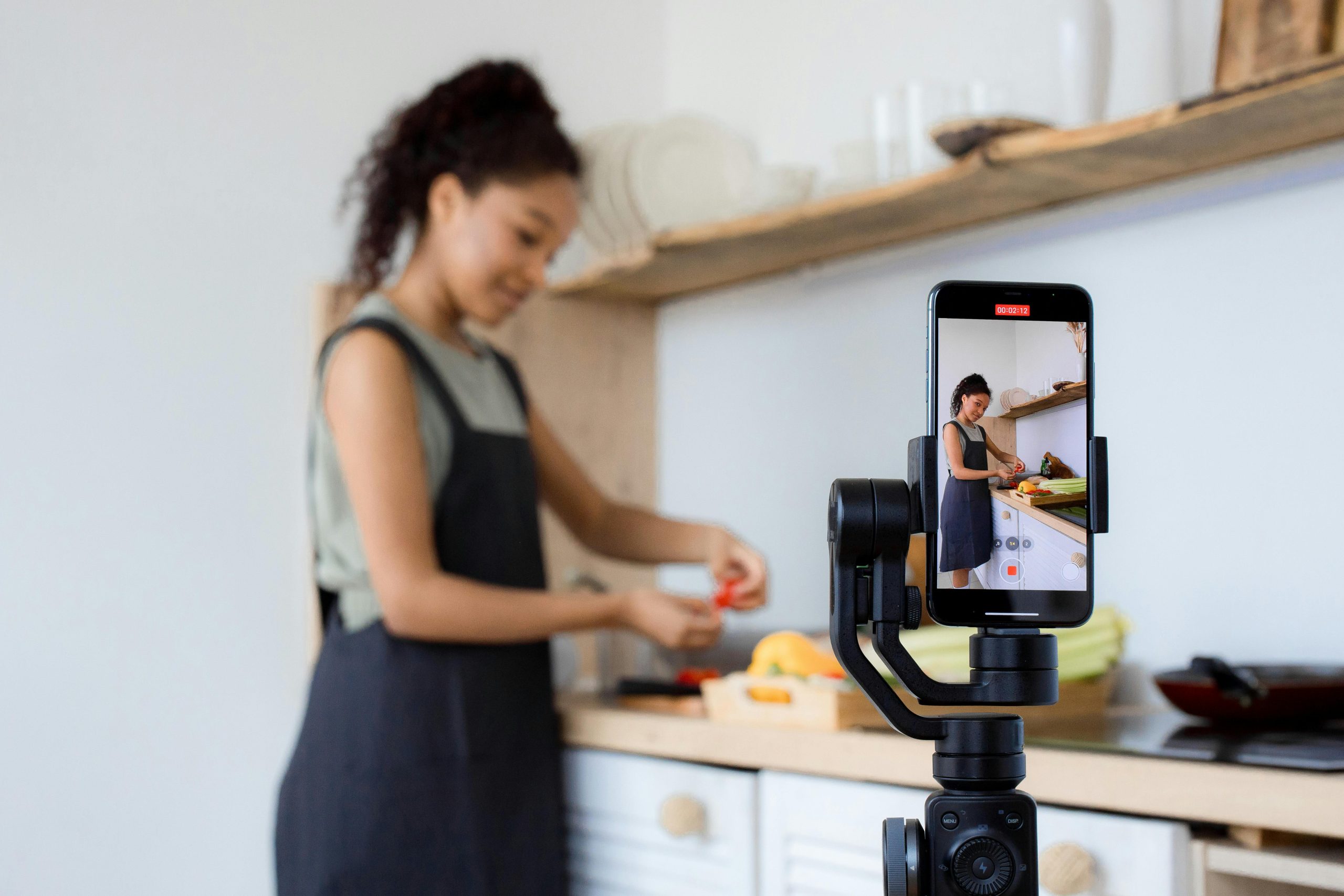 A woman records a cooking tutorial with her smartphone in a modern kitchen setting.